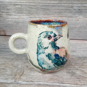 May include: A ceramic mug with a speckled cream-colored glaze and a rounded handle. The exterior features a hand-painted illustration of a bird with blue, green, and peach plumage. The inner rim has a vibrant, glossy blue and brown drip glaze finish.