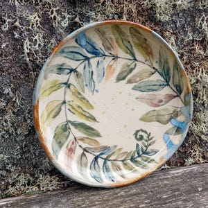 May include: A round ceramic bowl with a cream-coloured base featuring a hand-painted botanical design of fern leaves in shades of blue, green, and brown. The rim of the bowl has a warm orange-brown glaze, and the interior surface is speckled.