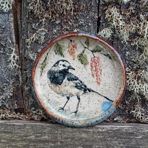 Puede incluir: Un plato de cerámica redondo con una lavandera blanca pintada a mano con plumaje negro y blanco. El fondo del plato es de color crema moteado con hojas verdes y amentos de color marrón rojizo que cuelgan de una rama sobre el ave.