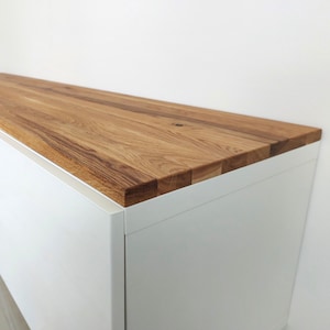 May include: A rectangular cabinet featuring a smooth, white matte finish on the sides and front, topped with a thick, natural wood surface. The wood top displays a prominent grain pattern with varied brown tones and visible joinery along the edges.