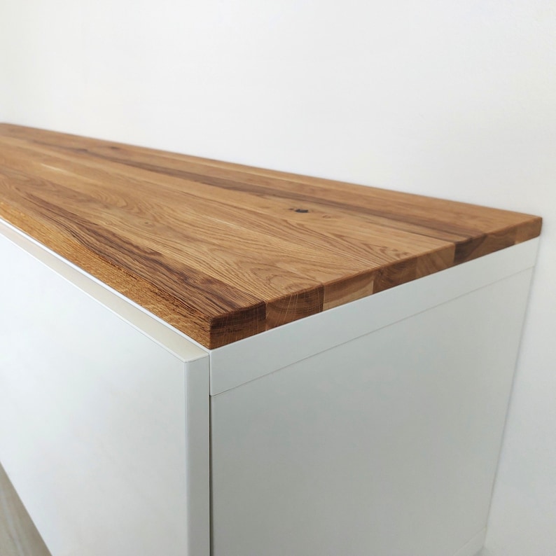 May include: A rectangular cabinet featuring a smooth, white matte finish on the sides and front, topped with a thick, natural wood surface. The wood top displays a prominent grain pattern with varied brown tones and visible joinery along the edges.