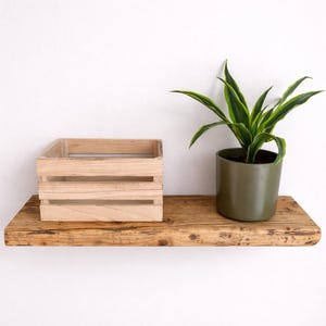 Peut inclure: Une &eacute;tag&egrave;re flottante rectangulaire en bois avec une finition &agrave; grain naturel. &Agrave; gauche se trouve une petite caisse en bois clair &agrave; lattes horizontales. &Agrave; droite, un pot cylindrique vert olive fonc&eacute; contient une plante verte et jaune.