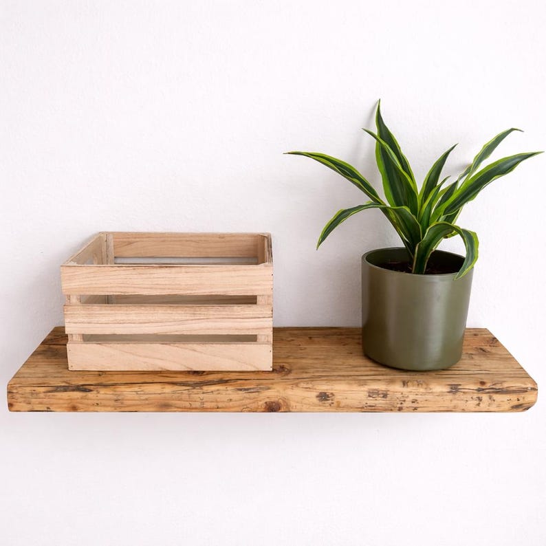 Peut inclure: Une &eacute;tag&egrave;re flottante rectangulaire en bois avec une finition &agrave; grain naturel. &Agrave; gauche se trouve une petite caisse en bois clair &agrave; lattes horizontales. &Agrave; droite, un pot cylindrique vert olive fonc&eacute; contient une plante verte et jaune.