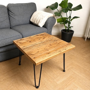 Könnte beinhalten: Quadratischer Couchtisch mit einer hellbraunen Tischplatte aus Altholz und schwarzen Haarnadelbeinen. Der Tisch steht vor einem grauen Sofa und einer Topfpflanze. Der Tisch ist ca. 60 cm breit und 46 cm hoch.