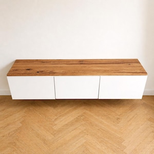 May include: A wall-mounted floating cabinet featuring a natural wood grain top surface and three smooth, solid white rectangular front panels. The unit has a horizontal rectangular shape with clean lines and a flat top, providing a storage solution.
