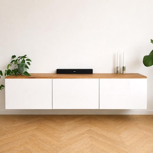 May include: A wall-mounted floating cabinet featuring three white rectangular doors and a natural wood top surface. A black soundbar is centered on the wood top, flanked by a green potted plant on the left and two tall candles on the right.