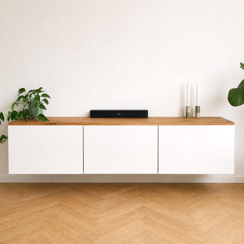 May include: A wall-mounted floating cabinet featuring three white rectangular doors and a natural wood top surface. A black soundbar is centered on the wood top, flanked by a green potted plant on the left and two tall candles on the right.