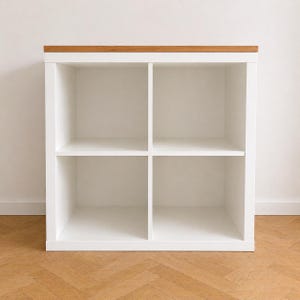 May include: A square white storage unit featuring four open cubby compartments arranged in a two-by-two grid. The unit is topped with a flat, natural light brown wooden surface that spans the entire width of the white frame.