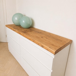 May include: A white six-drawer chest of drawers with a natural wood top. Two light green glass spheres sit on the top. The chest of drawers is in a room with white walls and a light-coloured wood floor.