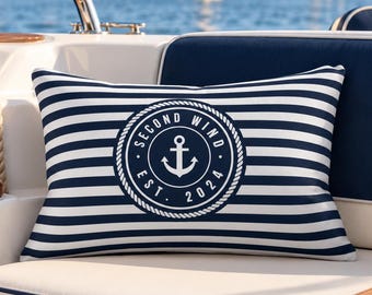 Könnte beinhalten: Ein rechteckiges Zierkissen mit horizontalen Streifen in Marineblau und Weiß. In der Mitte befindet sich ein kreisförmiges Emblem mit Seilrand, einem Ankersymbol sowie dem Text "SECOND WIND" und "EST. 2024" in weißer Schrift.