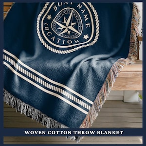 May include: A navy blue woven cotton throw blanket with a white nautical compass design and rope border. The blanket has white stripes and fringe. The text "BOAT NAME LOCATION" is visible on the blanket.