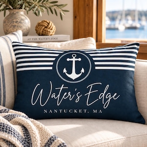 May include: A rectangular navy blue throw pillow featuring a white horizontal striped pattern at the top, a central white anchor icon inside a circular rope border, and the white text Water's Edge followed by NANTUCKET, MA below the anchor.