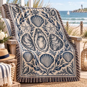Op de afbeelding: Een geweven plaid met een gedetailleerd marineblauw en crèmekleurig patroon van diverse schelpen, waaronder oesters, sint-jakobsschelpen en zanddollars, afgewisseld met zeewiermotieven. De deken is afgewerkt met decoratieve franjes aan de randen.