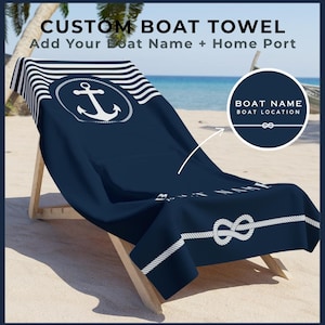 Op de afbeelding: Marineblauwe, op maat gemaakte boothanddoek met witte nautische designelementen, waaronder een anker, een touwknoop en strepen. De handdoek is gedrapeerd over een houten strandstoel op een zandstrand. De tekst op de handdoek luidt "CUSTOM BOAT TOWEL" en "Add Your Boat Name + Home Port".