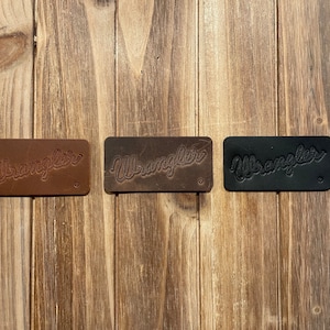 May include: Three leather patches with the word "Wrangler" embossed in a script font. The patches are brown, dark brown, and black.