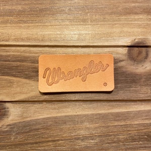 May include: A brown leather patch with the word "Wrangler" embossed in a rope design. The letter "R" in Wrangler is circled with a small "R" inside the circle.