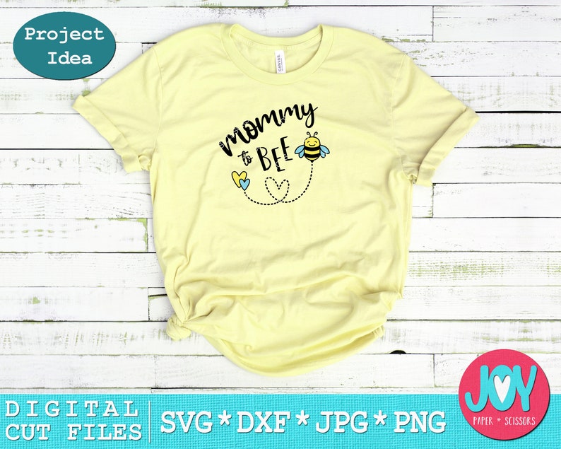 May include: A yellow t-shirt with the text "mommy to bee" in black lettering. A small bee with black and yellow stripes and blue wings is above the text. Two blue and yellow hearts are below the text with a dotted line connecting them to the bee.