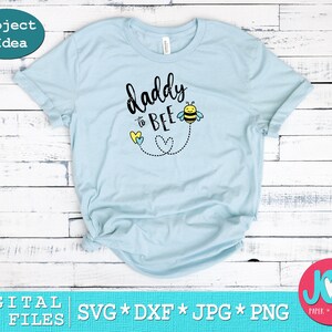 May include: Light blue t-shirt with the text "daddy to bee" in black lettering. A cartoon bee with yellow and black stripes is flying above the text. Two yellow and blue hearts are below the text.