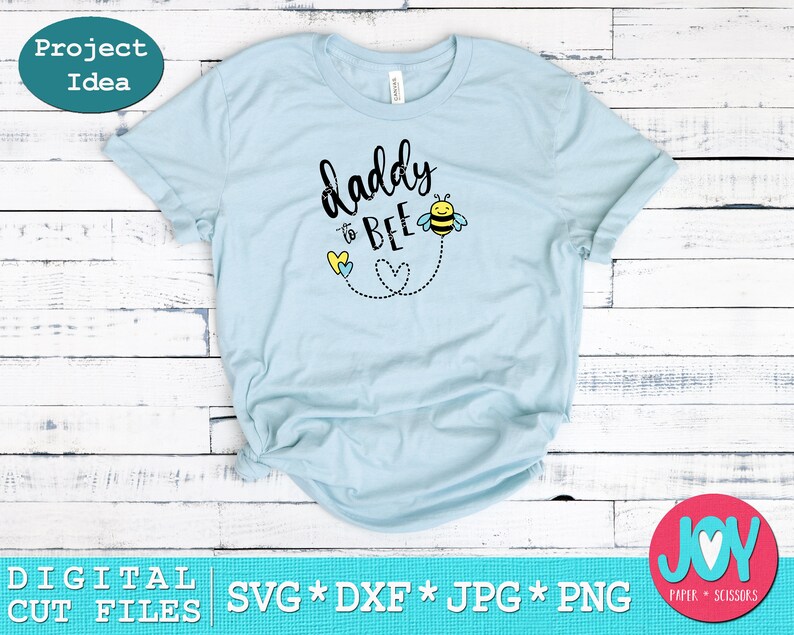 May include: Light blue t-shirt with the text "daddy to bee" in black lettering. A cartoon bee with yellow and black stripes is flying above the text. Two yellow and blue hearts are below the text.