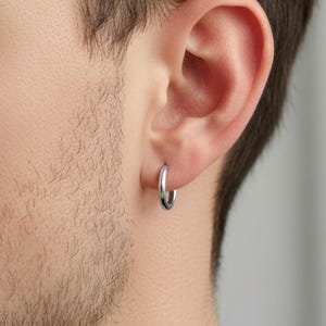May include: A close-up view of a person's ear with a small, silver hoop earring. The earring is circular and shiny, reflecting light. The person has short, dark hair and a beard.