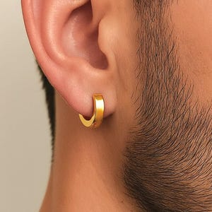 May include: A close-up of a gold hoop earring. The earring is a small, thick band that curves around the earlobe. The earring has a simple, classic design. The background is a blurred image of a person's ear and face.