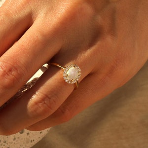 Könnte beinhalten: Ein goldener Ring mit einem großen ovalen weißen Opal-Stein, der von einem Heiligenschein aus kleineren klaren Steinen umgeben ist.