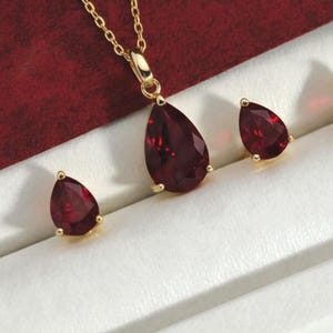 May include: A gold-toned jewelry set featuring a pendant necklace and matching earrings. The pendant and earrings showcase teardrop-shaped, deep red gemstones. The set is displayed in a white jewelry box with a red velvet lining.