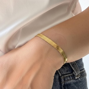 May include: A gold-toned herringbone snake chain bracelet featuring a flat, interlocking chevron pattern. The metallic jewelry piece rests on a wrist, showcasing a polished finish and a flexible, woven design that lies flat against the skin.
