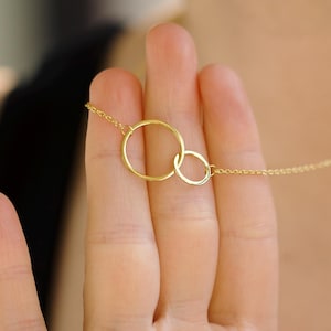 May include: A delicate gold necklace featuring two interlocking circles. The circles are connected by a fine chain, creating a minimalist and elegant design. The necklace is shown against a blurred background, highlighting the jewelry's simplicity and shine.