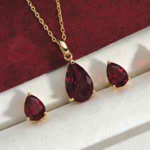 May include: A gold-toned jewelry set featuring a pendant necklace and matching earrings. The pendant and earrings showcase teardrop-shaped, deep red gemstones. The set is presented in a white jewelry box with a red velvet lining.