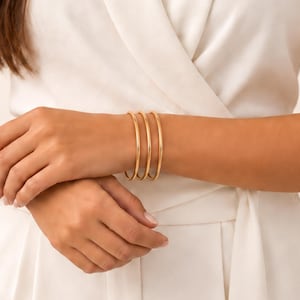 Puede incluir: Tres brazaletes finos y lisos de metal en tono dorado que se llevan en la muñeca. Las pulseras presentan un diseño circular sencillo con un acabado pulido y reflectante. Cada brazalete es idéntico en tamaño y forma, creando un efecto apilado.