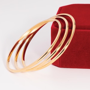 May include: Four gold-colored bangles are displayed on a white surface, with a red velvet box in the background. The bangles are thin and circular, reflecting light. The jewelry is simple and elegant.