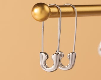 May include: Close-up of silver safety pin earrings hanging on a gold display stand. The earrings have a unique design, resembling safety pins. The background is a warm, neutral color, highlighting the jewelry.