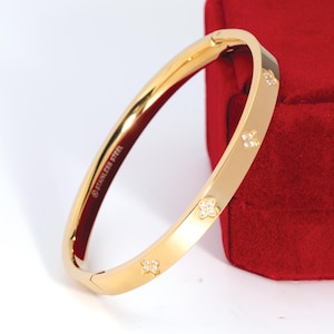 May include: A gold-colored stainless steel bangle bracelet with four small, sparkling flower-shaped embellishments. The bracelet is hinged and rests against a red velvet box. The words "STAINLESS STEEL" are printed on the inside of the bracelet.