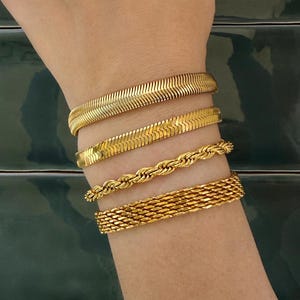 Puede incluir: Un conjunto de cuatro pulseras doradas en la muñeca. La colección incluye dos cadenas planas de espiga, una cadena de cuerda trenzada y una cadena de malla ancha, todas con un acabado metálico pulido y un color oro amarillo consistente.