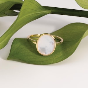 May include: A gold ring featuring a smooth, oval-shaped white mother-of-pearl inlay set within a polished gold bezel. The band is thin and circular, showcasing a clean, metallic finish that complements the iridescent, light-toned stone center.