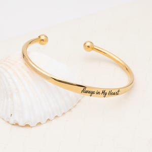 Könnte beinhalten: Ein goldfarbenes Metallarmband in Manschettenform mit polierter Oberfläche und zwei kugelförmigen Endkappen. Die Außenseite des Armbands ist mit dem schwarzen kursiven Text Always in My Heart entlang des gebogenen Bandes graviert.