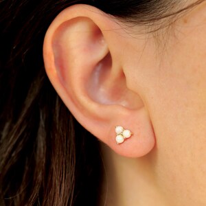 May include: A gold stud earring with three small, white, round pearls. The earring is set on a person's ear, showcasing the delicate design and subtle elegance of the jewelry.