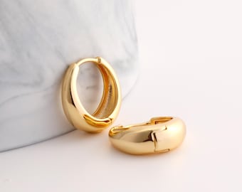May include: A pair of gold-toned hoop earrings with a thick, rounded, and polished surface. One earring is positioned upright, while the other lies flat, showcasing the smooth, reflective metal finish and the secure hinged clasp mechanism.