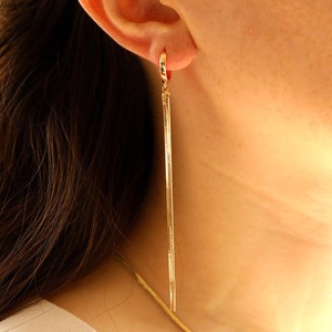 Puede incluir: Un par de aretes largos y delgados de oro que cuelgan del lóbulo de la oreja. Los aretes están hechos de una cadena delicada.