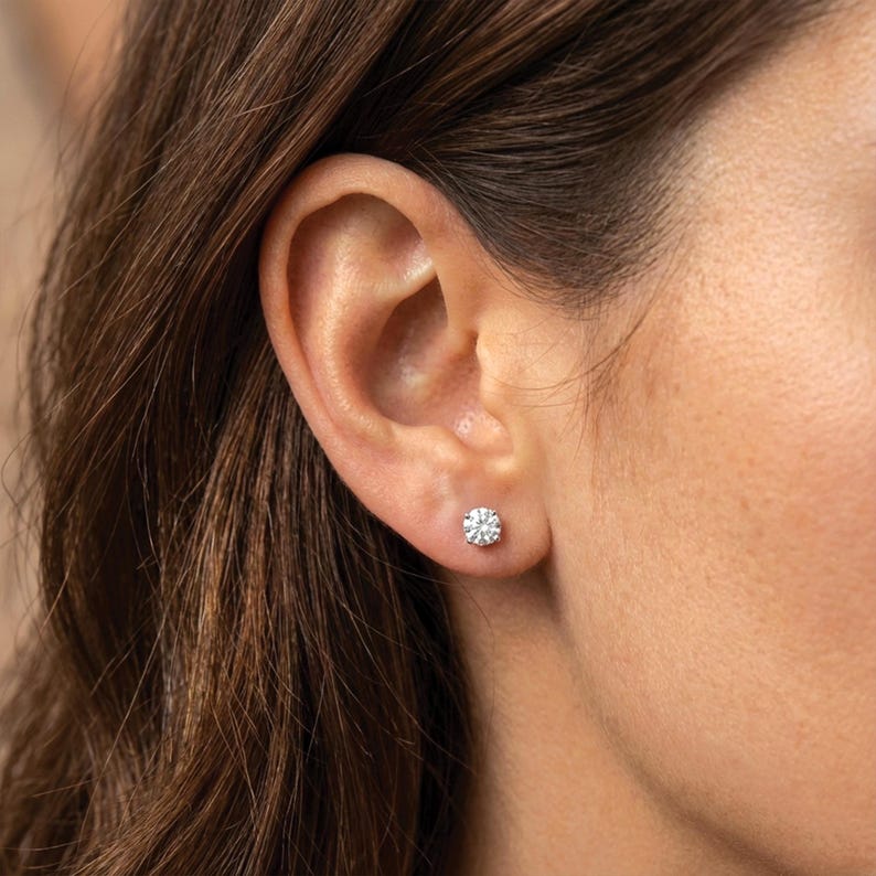 K&ouml;nnte beinhalten: Nahaufnahme eines Ohrs einer Frau mit einem einzelnen, runden, funkelnden Diamant-Ohrstecker. Der Ohrring ist in einem einfachen, klassischen Design gehalten und reflektiert das Licht. Die Haut der Frau ist hell und ihr Haar ist braun.