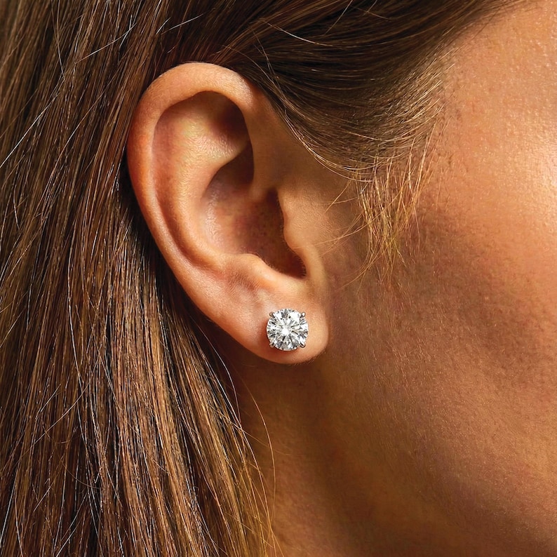 K&ouml;nnte beinhalten: Nahaufnahme eines funkelnden Diamantohrsteckers. Der rund geschliffene Diamant ist in einer wei&szlig;en Metalleinfassung gefasst und wird an einem Ohr getragen. Der Ohrring reflektiert das Licht und erzeugt ein brillantes Funkeln.
