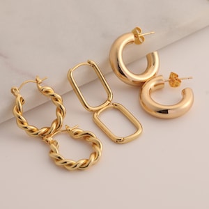 May include: A collection of gold-colored earrings in various styles. The earrings include twisted hoops, rectangular hoops, and chunky, rounded hoops. The earrings are displayed on a light-colored surface, showcasing their polished finish and design.