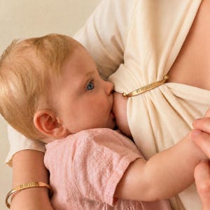 May include: A gold-toned metal nursing clip and a matching gold-toned metal bracelet, both engraved with the date 01.10.2025. The nursing clip features small spherical ends and is used to secure a cream-colored fabric garment.
