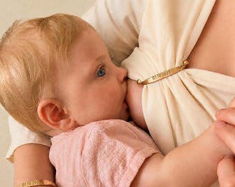 May include: A gold-toned metal nursing clip and a matching gold-toned metal bracelet, both engraved with the date 01.10.2025. The nursing clip features small spherical ends and is used to secure a cream-colored fabric garment.