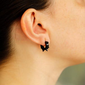 May include: Black hoop earrings with a spiked design. The earrings are made of a dark material and feature multiple small spikes around the hoop. The earrings are shown on an ear.