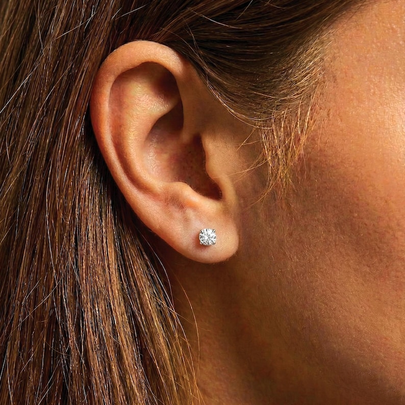 K&ouml;nnte beinhalten: Nahaufnahme eines Ohrs mit einem einzelnen, runden Diamant-Ohrstecker. Der Ohrring funkelt, gefasst in einem klassischen Design. Die Person hat lange, hellbraune Haare. Ein zeitloses Schmuckst&uuml;ck.