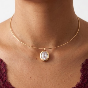 May include: A gold necklace featuring a clear, oval pendant. The pendant is set in a gold bezel and suspended from a fine, gold chain. The necklace is worn around the neck.