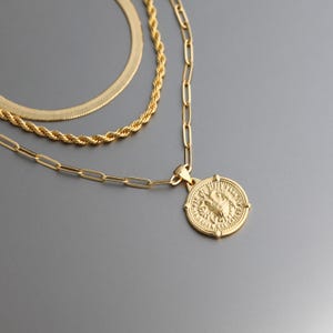 May include: A gold-coloured layered necklace set. The set includes a herringbone chain, a rope chain, and a chain with rectangular links. The lowest chain features a round pendant with a detailed design.