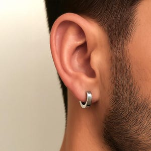 May include: A close-up of a person's ear with a small, silver hoop earring. The earring is a simple, minimalist design, reflecting light. The skin tone is light, and the background is a neutral color.